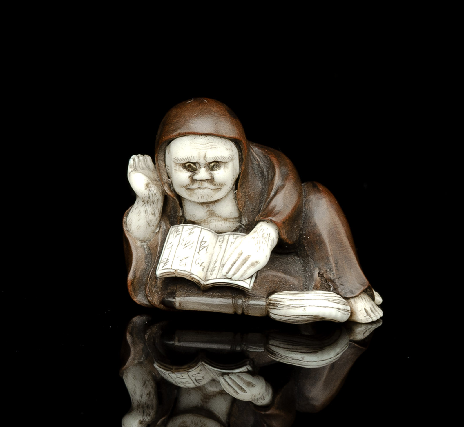 <b>Netsuke des Daruma ein Buch lesend mit Hossu-Wedel aus Holz teils mit Elfenbein und Horn eingelegt</b>