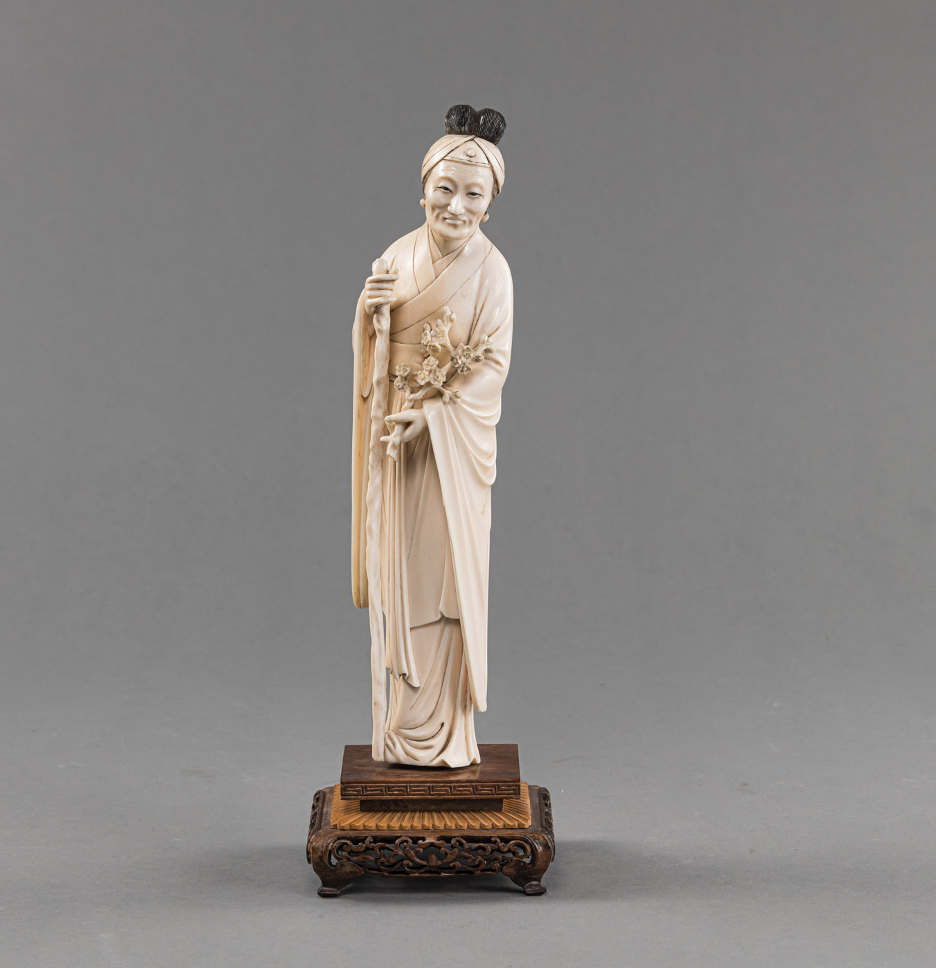 <b>Stehende Elfenbein-Figur einer älteren Dame in langem Gewand mit Gehstock und Pflaumenblütenzweig, montiert auf geschnitztem Holzsockel</b>