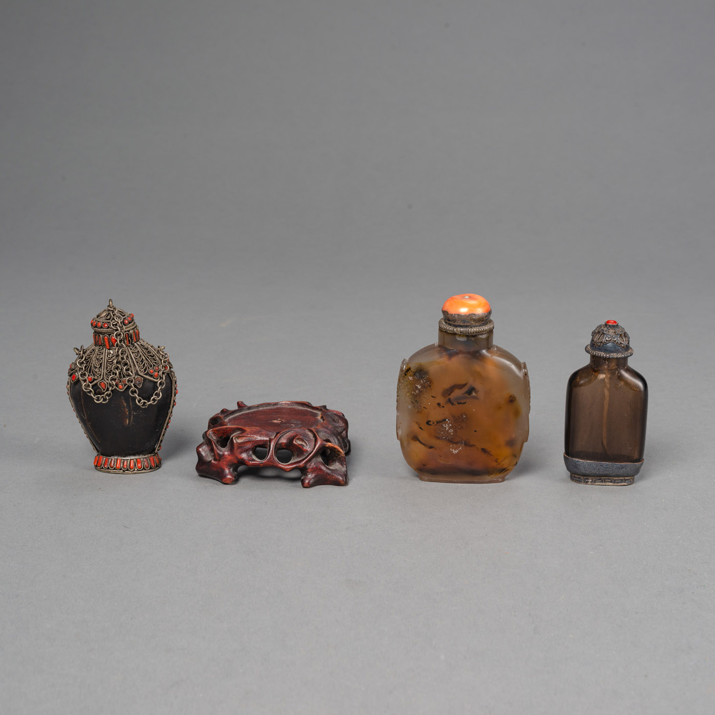 <b>THREE SNUFF BOTTLES, PARTLY METAL-FITTED, OF AGATE, WITH CORAL-INLAID STOPPERS AND FITTINGS, AND A WOOD STAND</b>
