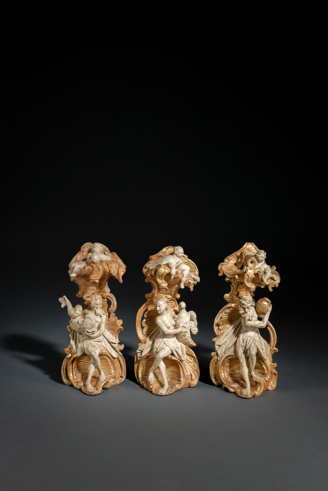 The three Mythological figures with their attributes carved in full round and sitting in front of a large volute surmounted by a rocaille tooled baldachin each on topped by a putto with a fish, a swan and a water vessel. Limewood, white polychromy and gilding. Remnants of old polychromy with retouches, minor restorations and traces of age. With later carved limewood wall bases, gilt and with white polychromy.